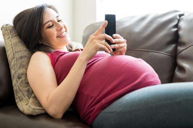 Pregnant laying on couch looking at pregnancy app on cell phone.